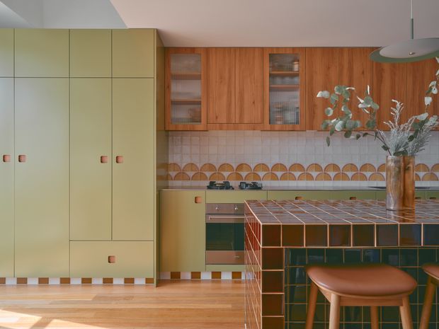 A warm, retro-inspired kitchen with pale olive-green full-height cabinetry, rich timber wall cabinets, a tiled backsplash with a geometric scalloped border, and a tiled island or peninsula in dark green and terracotta tones. A leather-topped stool and pendant light reinforce the mid-century character, while timber flooring and a vase of foliage add warmth.