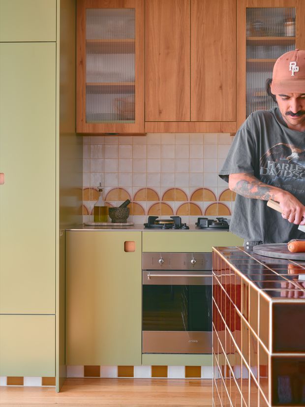 A warm, retro-inspired kitchen with olive-green cabinetry, timber overhead cupboards, ribbed glass-front doors, a tiled splashback featuring a geometric scalloped accent, and stainless-steel appliances. A person is preparing food beside a dark tiled kitchen island or peninsula with copper-toned grout and framing. The room has timber flooring and a playful, highly individual colour and material palette.