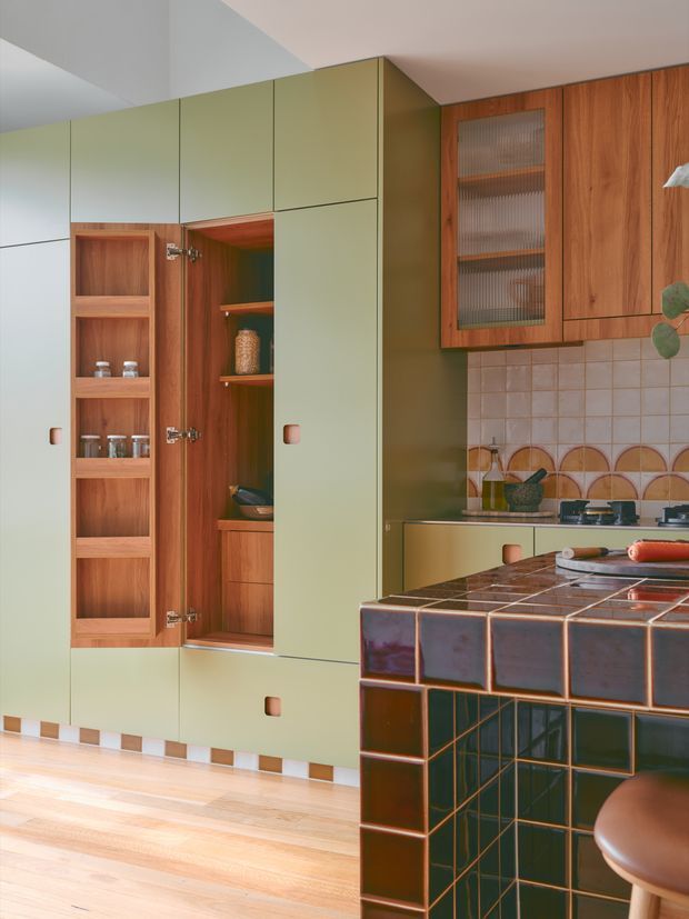 A detailed view of a colourful contemporary kitchen with floor-to-ceiling pale green cabinetry, integrated open shelving and warm timber interiors. The upper cabinetry combines timber doors with ribbed glass fronts, while the backsplash features square cream tiles with a decorative semicircular patterned band. A dark glossy tiled island or peninsula with contrasting grout dominates the foreground, paired with timber stools. Light timber flooring and a pale ceiling complete the warm, graphic interior.
