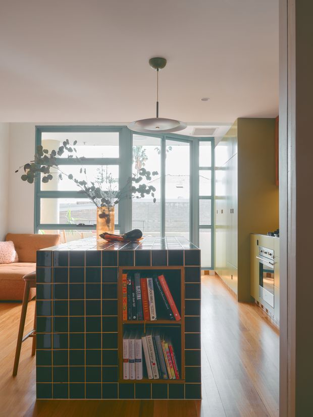 A warm, light-filled open-plan interior showing a compact kitchen and adjacent living area. The focal point is a dark teal tiled island with brass-toned grout and integrated open shelving filled with books. Behind it are pale timber floors, a muted green kitchen wall with a freestanding oven, extensive blue-grey framed glazing, and a peach-toned sofa. A simple suspended disc pendant hangs above the island, with a vase of branches and fruit bowl adding understated decor.