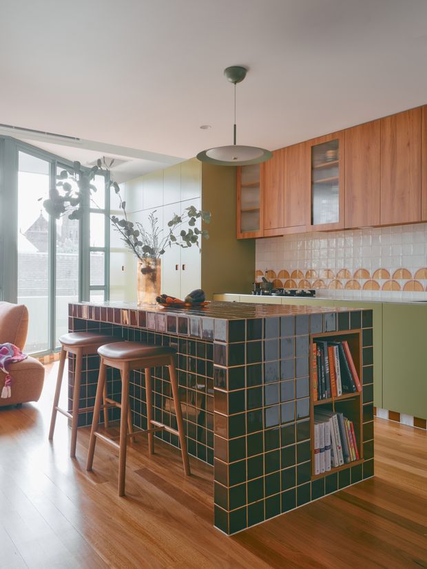 A bright, mid-century-inspired kitchen viewed from the living area, featuring a large dark green tiled island with timber edging and integrated open shelving, warm timber cabinetry, a patterned white tile splashback, timber flooring, and retro pendant lighting. Full-height glazing brings in abundant natural light, with a small seating area visible at left.
