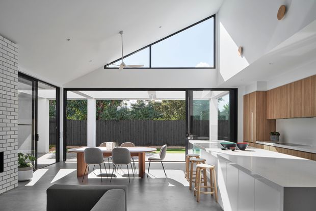 A bright, open-plan contemporary kitchen and dining area with a large white island, integrated timber cabinetry, dining table, stools, and generous black-framed glazing opening to a landscaped courtyard. A high triangular clerestory window, exposed ceiling fan, polished concrete floor, and white brick feature wall create an airy modern interior.
