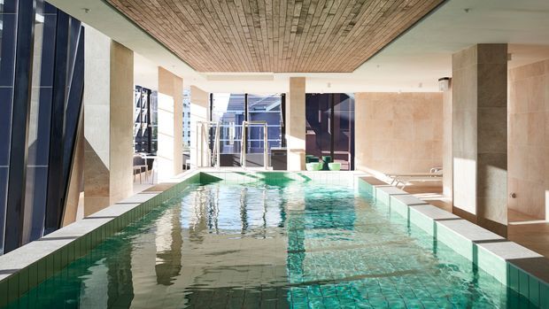 A bright indoor swimming pool beneath a large timber-lined ceiling, surrounded by pale stone or concrete columns and walls with extensive glazing. The pool has green-toned tiled edges and clear turquoise water, creating a refined modern aquatic interior.