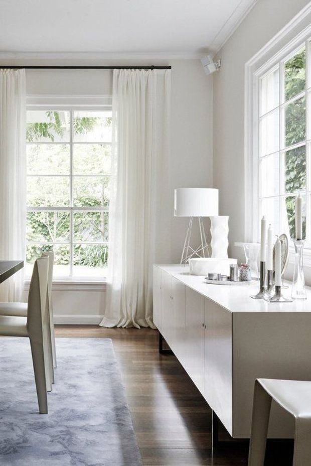 Bright, refined dining room with a long white built-in sideboard beneath large multi-pane windows. A white dining table and chairs, sheer floor-length curtains, contemporary table lamp, sculptural vase, and silver-toned tableware create a light monochromatic interior. Dark timber flooring and a pale area rug add contrast, while traditional window proportions and ceiling cornice details complement the contemporary furnishings.