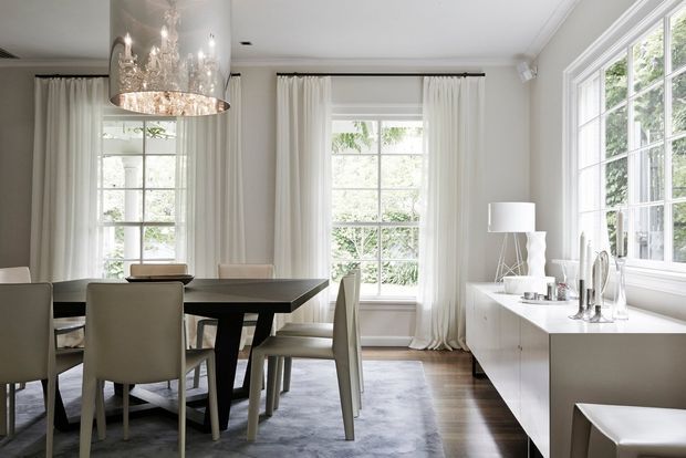 Bright, refined dining room with a large dark dining table, upholstered dining chairs, a dramatic crystal pendant light, full-height white curtains, and expansive multi-pane windows. A streamlined white sideboard with decorative objects and a table lamp runs along the right wall. Pale walls, dark timber flooring, and a soft grey area rug create a light contemporary-classic interior.