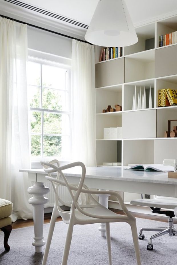 Bright, refined home office with a white desk spanning the room, sculptural white desk chairs, built-in open shelving and closed cabinetry, books and decorative objects, a large multi-pane window with sheer curtains, and a contemporary pendant light. The room has a predominantly white and pale neutral palette with abundant natural light.