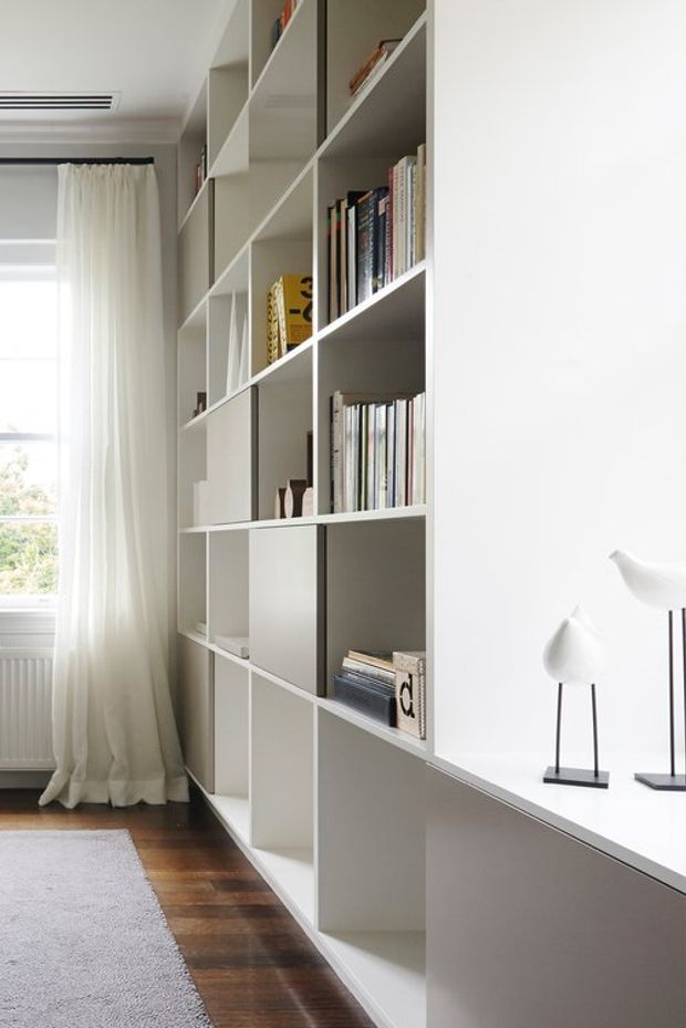 Bright, minimalist interior featuring a long wall of built-in white open shelving filled with books and decorative objects. A white built-in desk or cabinet runs along the right side, with modern sculptural lamps. The room has sheer floor-length curtains, a window, dark timber flooring, and a pale area rug.