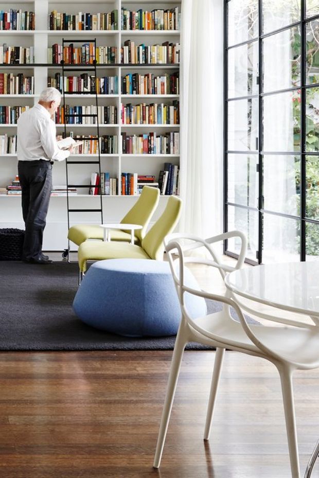 Bright contemporary living room with a full-height built-in white bookshelf filled with books, a black metal ladder rail, lime-green lounge chairs, a rounded blue ottoman, and white sculptural dining chairs around a glass-topped table. Large black-framed grid windows bring in abundant natural light, while dark carpeting contrasts with warm timber flooring.