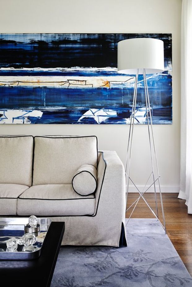 A refined living room featuring a light beige upholstered sofa with black piping, a tall white tripod floor lamp, a large abstract blue and white artwork, dark timber flooring, and a muted blue patterned rug. A black coffee table with a reflective tray and glassware sits in the foreground, while sheer white curtains frame the right side.