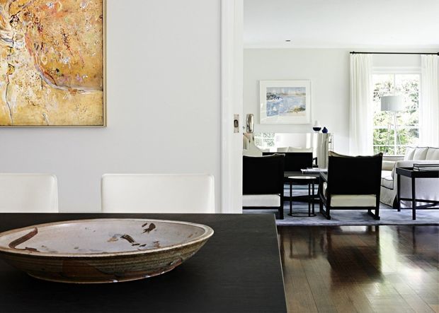 A refined open-plan dining and living area viewed from the dining table. The foreground features a dark dining table with a large handcrafted ceramic bowl and white upholstered dining chairs. Beyond it is a bright living room with dark armchairs, a pale sofa, side tables, artwork, and tall curtained windows. The space has light grey walls, dark timber flooring, and a restrained neutral palette with warm natural accents.