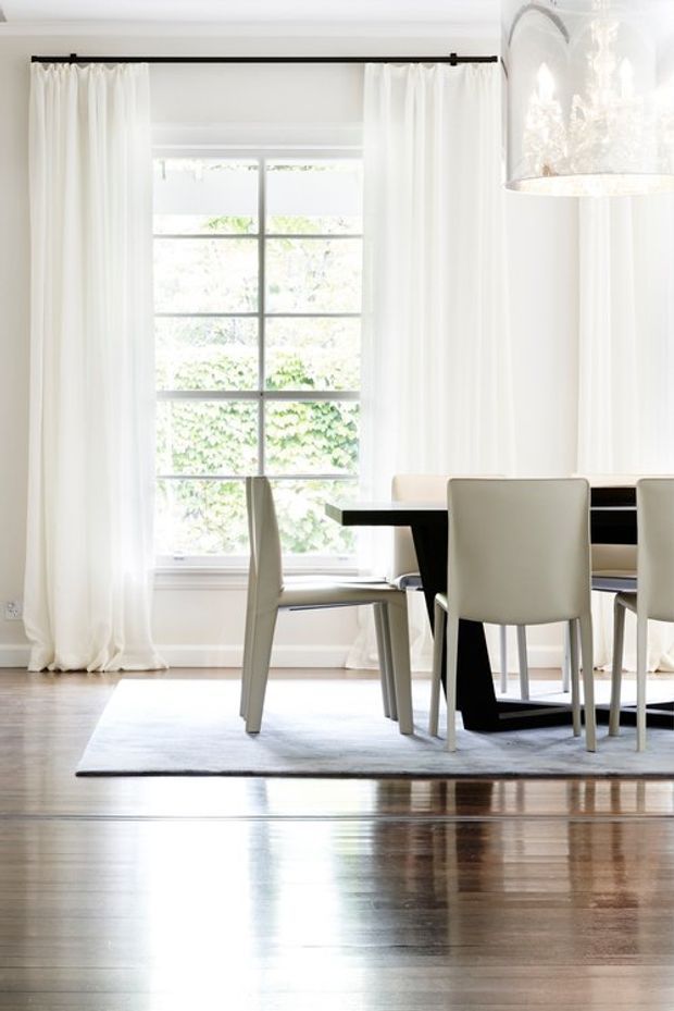 Bright, refined dining area with a black rectangular dining table and upholstered light dining chairs positioned on a pale rug. The room features tall divided-pane windows, full-length sheer white curtains, polished dark timber flooring, white walls and ceiling, and a contemporary pendant chandelier. Natural light creates a clean, airy atmosphere.