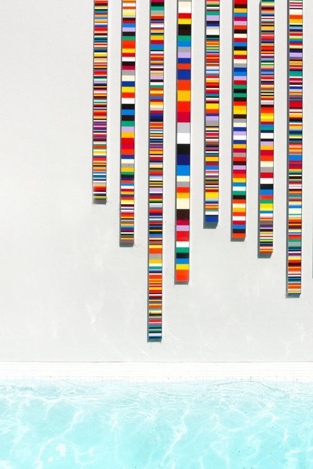 A minimalist architectural detail showing a white wall decorated with multiple vertical strips composed of tightly arranged, brightly coloured horizontal bands. The installation hangs above the edge of a turquoise swimming pool, creating a vivid, graphic contrast between the multicoloured artwork, white surface, and pale blue water.