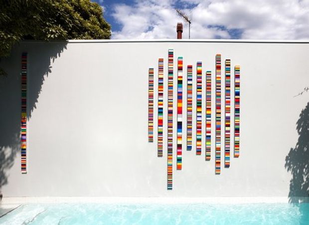 A white exterior wall beside a swimming pool, featuring a colourful vertical striped graphic or decorative installation arranged in a sound-wave-like pattern. A narrow matching stripe appears at the left edge. Trees and sky are visible above the wall, with strong sunlight and shadows.