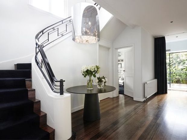 Bright, refined residential entry hall with a sweeping curved staircase, dark carpeted treads, polished dark timber flooring, white walls and trim, and a sculptural glass pendant light above a round console table. Black-framed detailing, a radiator, and full-height curtains frame the adjoining glazed opening.