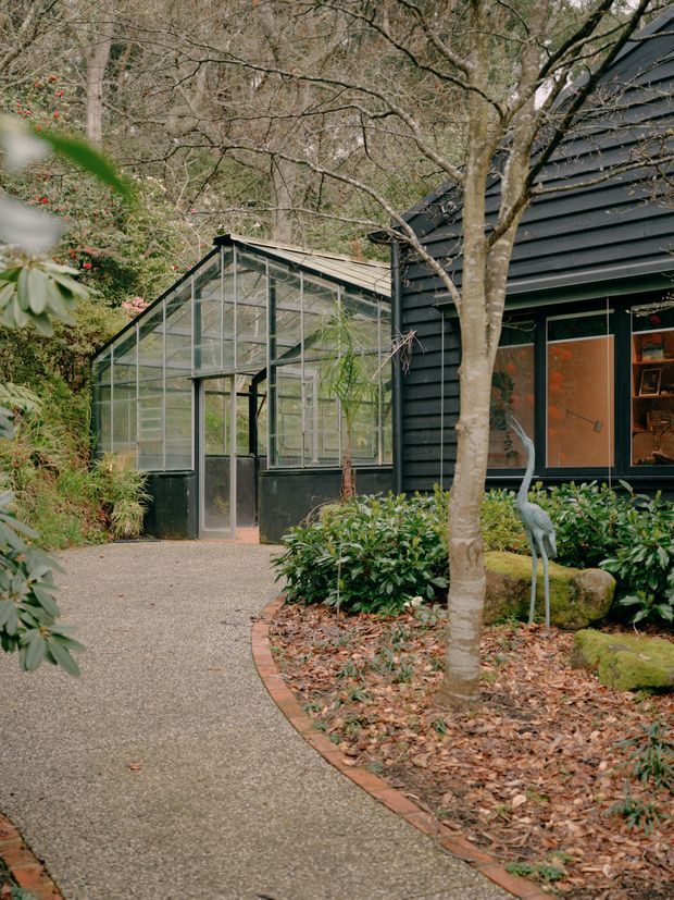 Exterior view of a dark-clad house set within a mature, leafy garden. A glazed greenhouse or conservatory with a pitched roof connects to the building beside a curved textured concrete walkway edged with brick pavers. Large windows reveal a warmly lit interior, while dense planting, moss-covered garden edging and a decorative heron figure create a natural woodland setting.