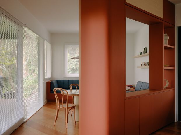 A warm, compact dining nook viewed through a richly terracotta-coloured built-in partition and display cabinetry. The room has pale timber flooring, extensive windows with slim blinds, a small dining table with curved timber chairs, and a deep teal upholstered bench. The cabinetry incorporates open shelving and a large internal opening that visually connects the spaces. The overall composition is contemporary with strong colour-blocking and natural light.