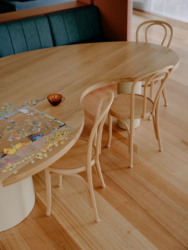 Warm dining area featuring a large light timber oval dining table with softly rounded edges, matching bentwood chairs, and a built-in teal upholstered bench seat. Light timber flooring, white walls, and a partially visible window create a bright, playful setting. A jigsaw puzzle and small amber glass vessel sit on the table.