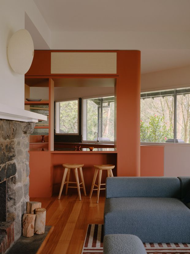 Warm, retro-inspired open-plan interior showing a compact kitchen opening into a living room. The kitchen features terracotta-orange painted cabinetry and structural framing, a dark benchtop, timber stools, timber flooring, and large windows with horizontal blinds. A substantial rough-stone fireplace with a white mantel occupies the left foreground, while a blue-grey sofa and patterned rug appear in the living area.