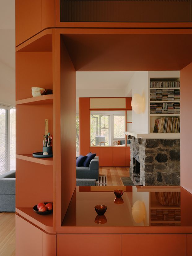Interior view through a warm terracotta built-in shelving and cabinetry composition, looking toward a living room and dining area. The foreground features open display shelves, rounded built-in detailing, glossy reflective cabinetry, and a deep orange-red finish. In the background are a blue-grey sofa, timber flooring, large windows and doors, a white wall, extensive book shelving, and a substantial exposed dark stone fireplace. The image has a highly curated, warm, retro-modern character with strong geometric framing and reflective surfaces.