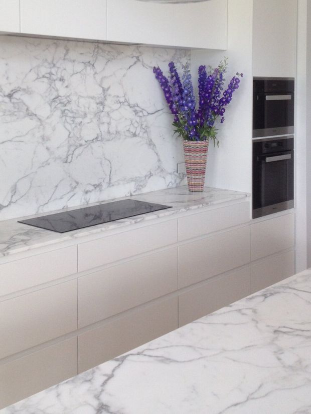 Bright contemporary kitchen with full-height white marble-look veined splashback and matching benchtops, handleless pale cabinetry, a black induction cooktop, and stacked black built-in ovens. A striped vase of purple flowers provides the main decorative accent.