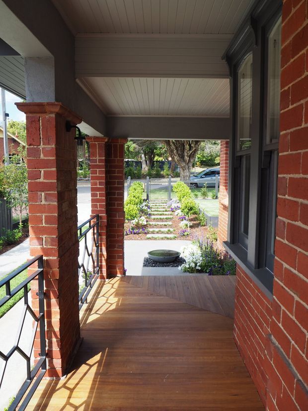 A covered front veranda with warm timber decking, red brick columns and walls, dark-framed windows, and a white-lined ceiling. Black metal railings border the elevated porch, which overlooks a landscaped front garden with stepping stones, shrubs, and flowers.