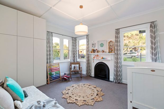 Bright, cozy children's bedroom with pale built-in cabinetry, a bed with colorful cushions, a traditional decorative fireplace, patterned curtains, soft grey carpet, and playful toys and artwork. Large windows provide abundant natural light, while a white pendant light and ornate ceiling add character.