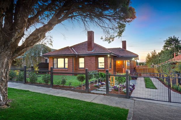 Street-facing exterior photograph of a single-storey red-brick bungalow with a terracotta tiled hipped roof, prominent brick chimneys, timber-framed windows, and a covered entry porch. The property has a landscaped front garden, low metal fencing and gate, paved paths, and mature trees.