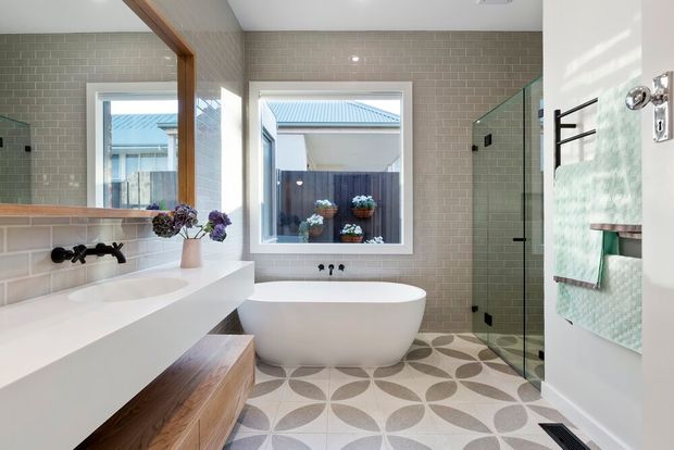 Bright contemporary bathroom with a freestanding white bathtub beneath a window, a long white double vanity with timber cabinetry, black wall-mounted tapware, pale subway-tile walls, patterned geometric floor tiles, and a frameless glass shower enclosure. A large mirror and folded towels are visible, with an outdoor courtyard seen through the window.