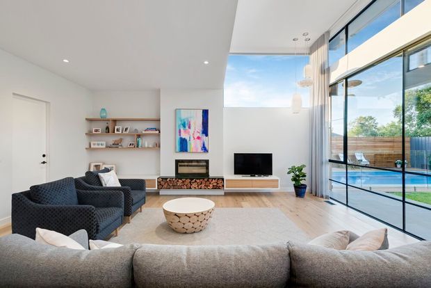 Bright contemporary living room with a high ceiling and expansive floor-to-ceiling glazing overlooking a pool and outdoor area. The room features a large sectional sofa, two dark armchairs, a round coffee table, built-in shelving, a linear fireplace with stacked firewood storage, television joinery, light timber flooring, and sheer curtains.