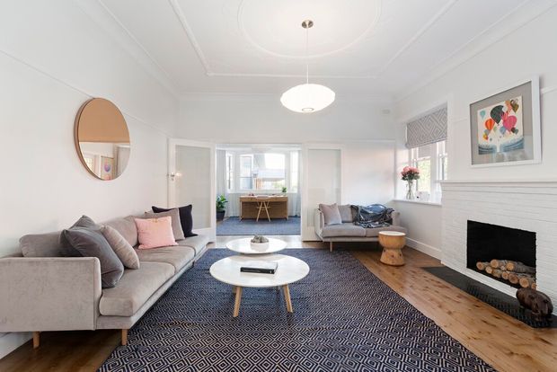 Bright, refined living room with white walls, decorative ceiling mouldings, a fireplace with white mantel surround, light timber flooring, and a large patterned navy rug. The room features a pale upholstered sectional sofa, armchair, round white coffee tables, a pendant light, framed artwork, and a view through to a dining or kitchen area.