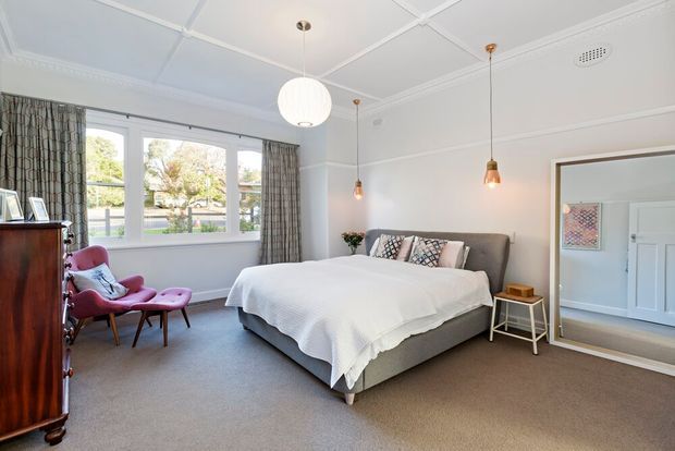 Bright, spacious bedroom with a contemporary upholstered bed, white bedding, patterned cushions, grey carpet, and large windows dressed with patterned curtains. The room includes a pink upholstered lounge chair, timber furniture, pendant lighting, white decorative ceiling detailing, and a large wall mirror reflecting a white door and artwork.