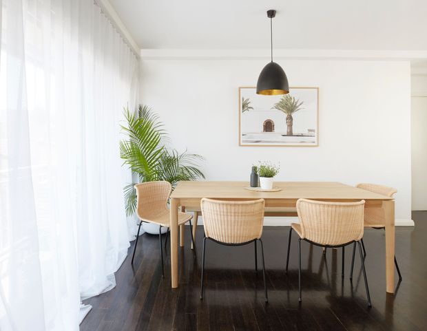 Bright dining room with a rectangular light timber dining table and woven rattan dining chairs, set against white walls and dark timber flooring. Sheer floor-to-ceiling curtains provide soft daylight, while a black pendant light, potted palm, and framed artwork add restrained decorative accents.
