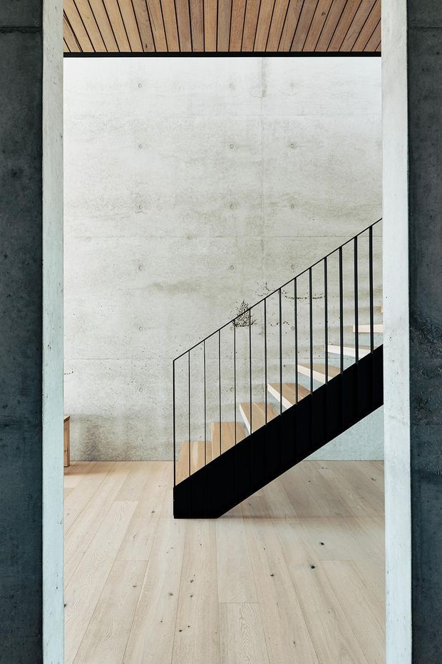 A minimalist interior stairway viewed through a concrete-framed opening. The space features a pale timber plank floor, a raw concrete wall with visible form-tie marks, a black metal stair balustrade and dark stair stringer, and a timber-lined ceiling. The composition emphasizes clean geometric lines, exposed materials, and natural light.