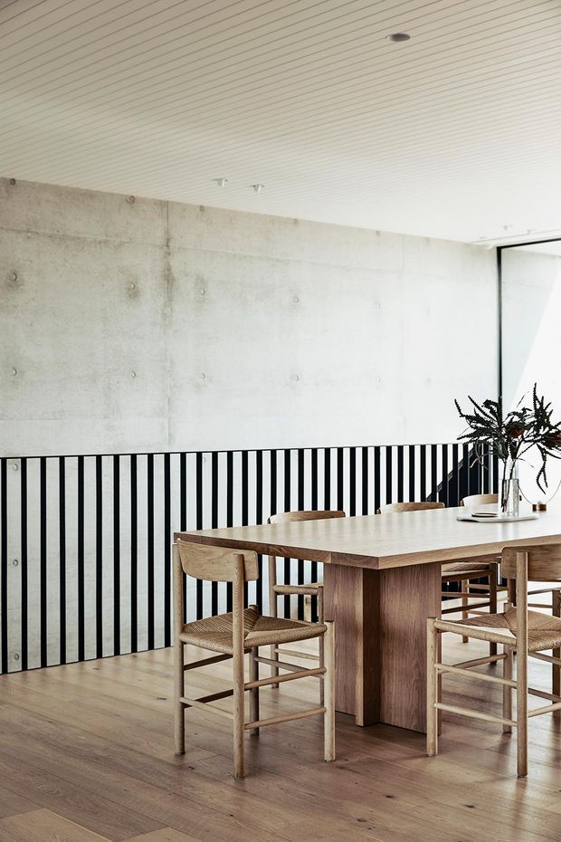 A bright, minimalist dining area with a large solid wood dining table and matching wooden chairs set against a raw concrete wall. A black vertical metal balustrade borders the space, while a white slatted ceiling, timber flooring, and full-height glazing contribute to the airy contemporary interior. A simple vase with foliage provides a natural accent.