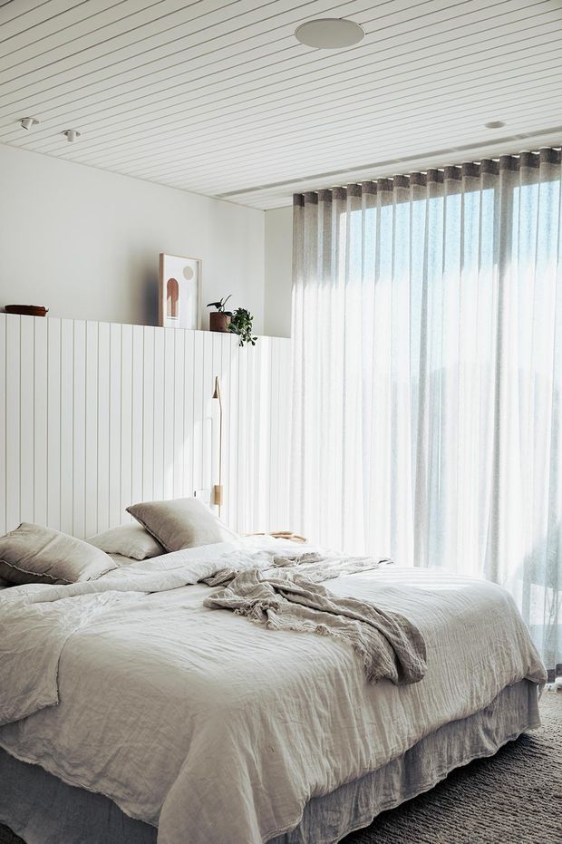 Bright, serene bedroom with a low white vertical-panel partition behind the bed, layered neutral linen bedding, sheer floor-to-ceiling curtains, a textured carpet, and a white slatted ceiling. Minimal artwork and a potted plant add subtle decorative detail.