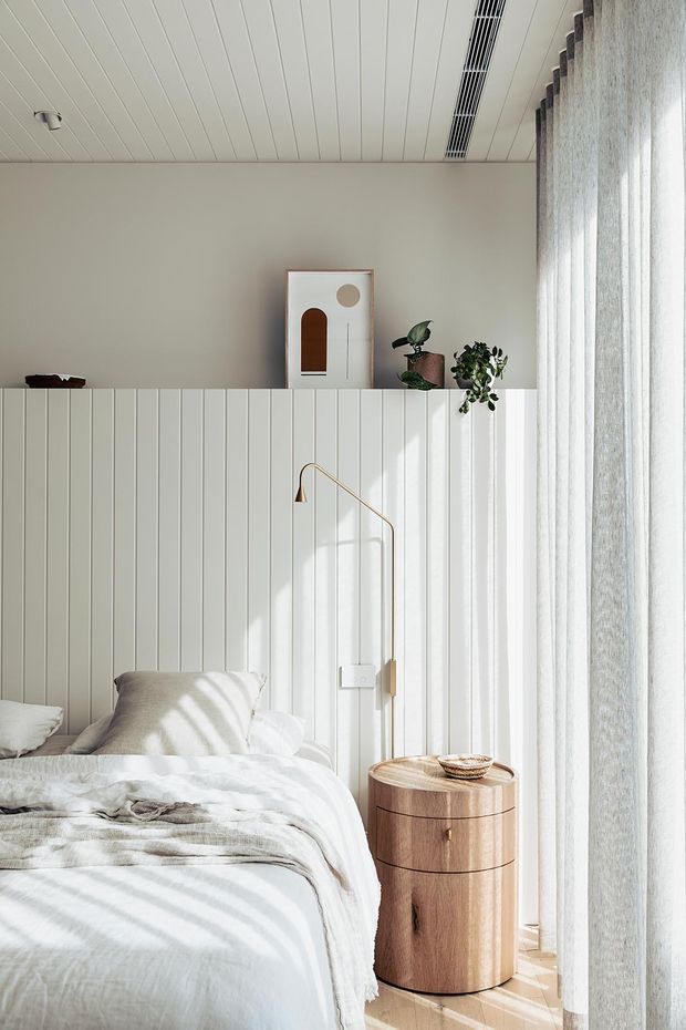 Bright, serene bedroom corner featuring a bed with layered white and grey bedding, a vertical white wall-panelled headboard, a slim brass wall-mounted reading light, and a rounded timber bedside table. Sheer full-height curtains filter abundant natural light, while potted plants and minimalist artwork add soft decorative accents. The white-lined ceiling includes recessed linear air conditioning or ventilation.