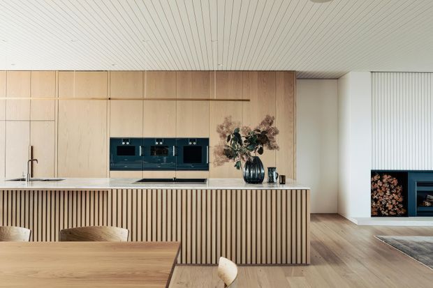 Contemporary open-plan kitchen and dining area with a long light-wood island featuring vertical slatted detailing, integrated dark appliances, pale timber cabinetry, and a matching linear timber ceiling. A timber dining table and chairs occupy the foreground, while a fireplace and media joinery are visible in the adjacent living area.