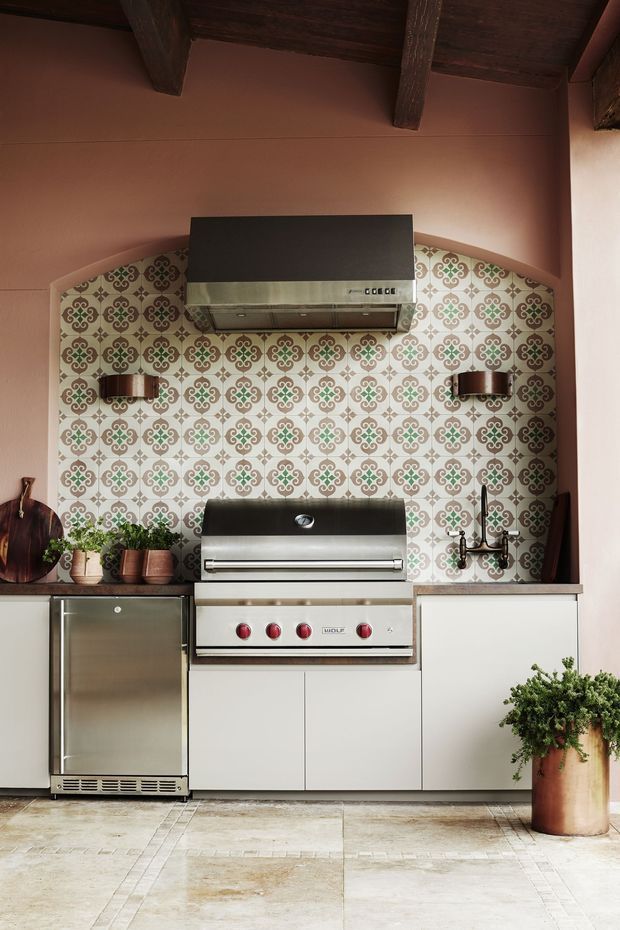 A compact built-in outdoor kitchen beneath a dark timber-beamed ceiling, featuring a stainless-steel barbecue, range hood, under-counter refrigerator, white cabinetry, and a decorative patterned tile splashback framed by warm pink plaster walls. Pale stone or porcelain paving covers the floor, with potted herbs and greenery adding an organic touch.
