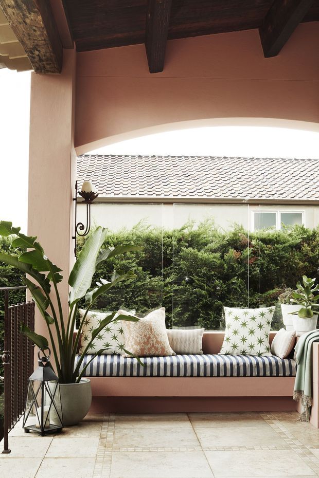 A covered outdoor terrace or courtyard lounge with a built-in cushioned bench beneath a dark timber-beamed ceiling. The space features warm terracotta plaster walls, pale stone or tile flooring, patterned cushions, tropical potted plants, a hanging lantern, and frameless glazing overlooking a planted courtyard and tiled roof.
