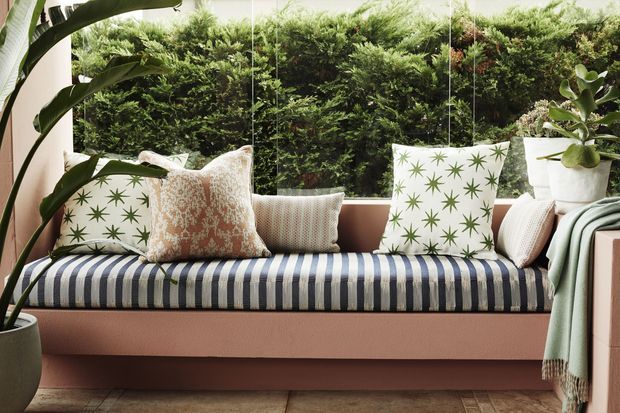 A bright, plant-filled sunroom or enclosed outdoor seating area with a built-in blush-pink bench beneath large glazing. The bench has a navy-and-white striped cushion, patterned decorative pillows, and a pale green throw, with lush greenery visible immediately outside. Large potted tropical plants frame the scene, and the floor appears to be warm-toned timber or tile.