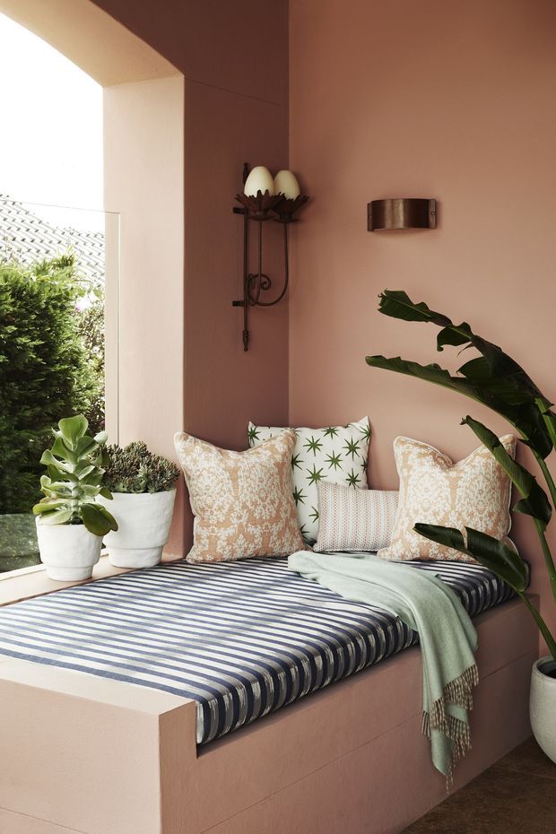 A warm, enclosed outdoor or semi-outdoor daybed nook with dusty rose plaster walls, a built-in upholstered platform, striped blue-and-white bedding, patterned cushions, a pale throw, potted plants, and a decorative wall sconce. Bright daylight enters from the open side, creating a relaxed, botanical retreat.