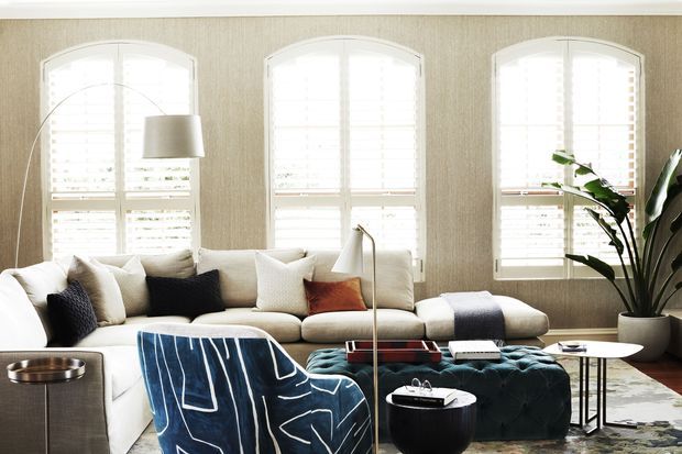 A bright, refined living room with a large cream sectional sofa arranged beneath three tall arched windows with white plantation shutters. The room includes a navy-and-white patterned accent chair, a deep teal tufted ottoman, a slim floor lamp, a round side table, a sculptural black coffee table, and a large indoor plant. Textured neutral wallcovering and layered furnishings create an inviting, contemporary-classic atmosphere.