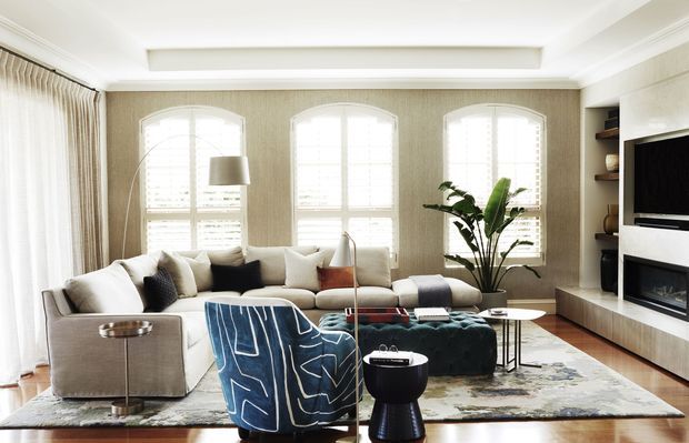 Bright, refined living room with a large sectional sofa arranged around a patterned ottoman and sculptural lounge chair. The room features tall arched windows with plantation shutters, textured neutral wallcovering, floor-to-ceiling curtains, timber flooring, built-in shelving, and a contemporary fireplace and television wall. A large indoor plant and layered rugs add warmth and texture.
