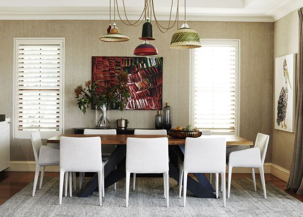 A refined dining room with a large dark rectangular dining table surrounded by upholstered white dining chairs. Textured neutral wallpaper, white plantation shutters, timber flooring, a pale area rug, colourful pendant lights, artwork, and a floral arrangement create a layered contemporary interior.
