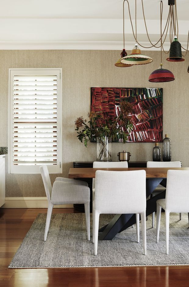 A refined dining room with a rectangular timber dining table, upholstered white dining chairs, and a large textured neutral rug over dark timber flooring. The room features textured beige wallcovering, white trim and plantation shutters, a colourful abstract artwork, decorative vases and dried botanical arrangements. A cluster of distinctive woven pendant lights hangs above the table.
