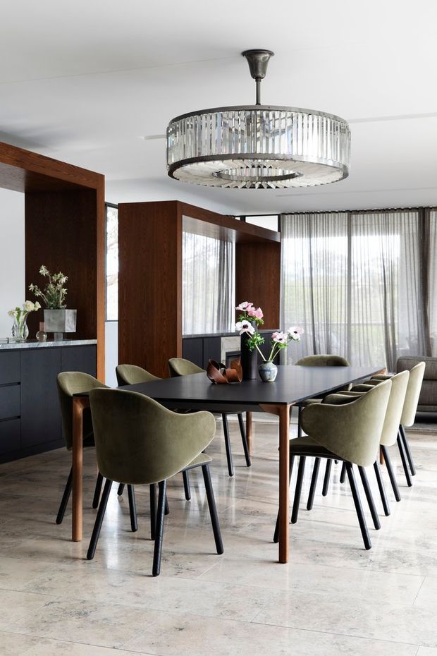A refined dining room with a large dark rectangular dining table surrounded by upholstered olive-grey dining chairs. The room features dark timber room dividers and cabinetry, full-height sheer curtains, pale stone-look flooring, and a prominent circular crystal pendant light. Floral arrangements and understated styling add an elegant, contemporary atmosphere.