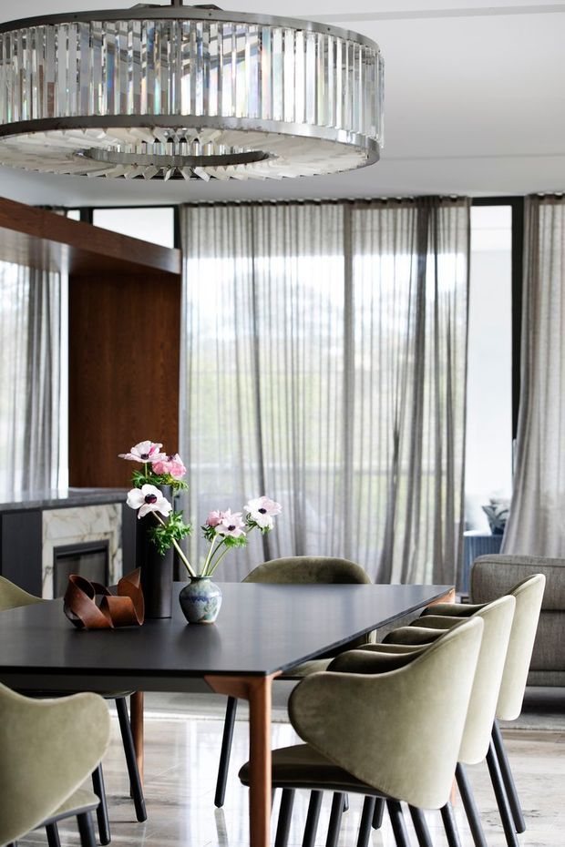 Elegant contemporary dining room with a large dark dining table surrounded by upholstered curved dining chairs. The room features floor-to-ceiling sheer curtains, expansive glazing, a marble-faced fireplace, dark timber wall panelling, pale flooring, and a prominent circular crystal-and-metal chandelier.