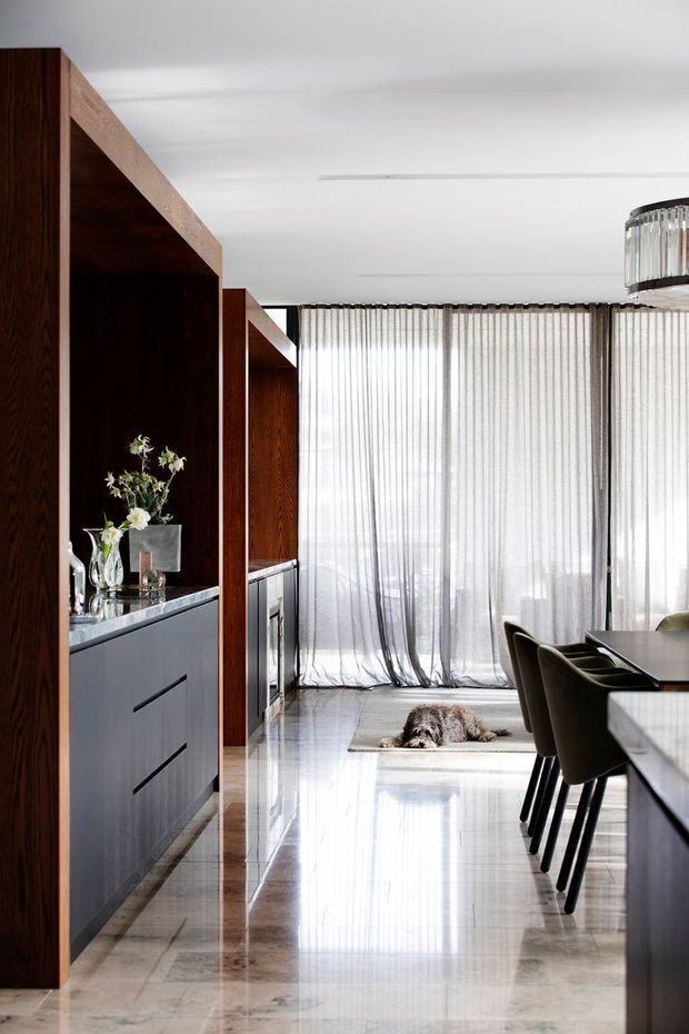 A refined open-plan dining and living area with dark timber built-in cabinetry, a grey stone-topped console, polished pale stone flooring, floor-to-ceiling sheer curtains, and a contemporary dining table with upholstered chairs. A sculptural pendant light hangs above the dining area, while a small dog rests on a rug near the glazed exterior.