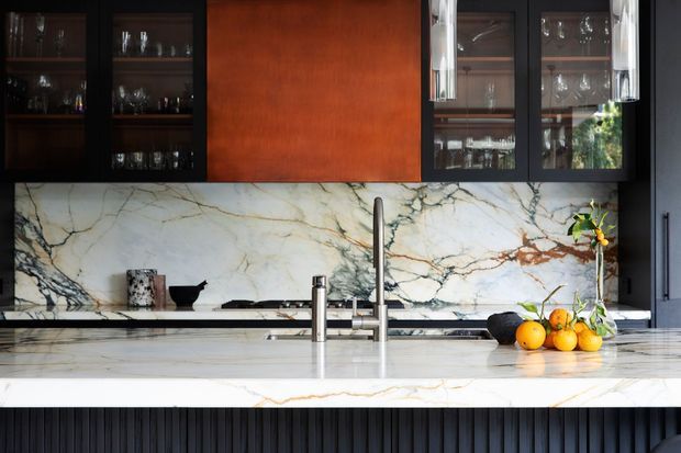 A refined contemporary kitchen featuring a large white marble island and matching dramatic veined marble splashback. Dark ribbed cabinetry and black-framed glass display cabinets contrast with a warm timber feature panel, while brass-toned pendant lights and a stainless-steel tap add detail. Fruit and small accessories sit on the island.