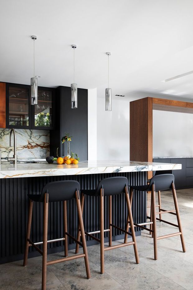 A sophisticated contemporary kitchen with a large marble island and integrated breakfast bar. The island features a deeply fluted black base and a white marble benchtop with prominent grey and brown veining, paired with four dark upholstered timber bar stools. Black built-in cabinetry with glazed display doors lines the rear wall, alongside a marble splashback. Three slim cylindrical pendant lights hang above the island. A timber-framed opening and dark cabinetry continue into the adjacent space. The room has a bright, refined, high-contrast palette with timber and stone accents.